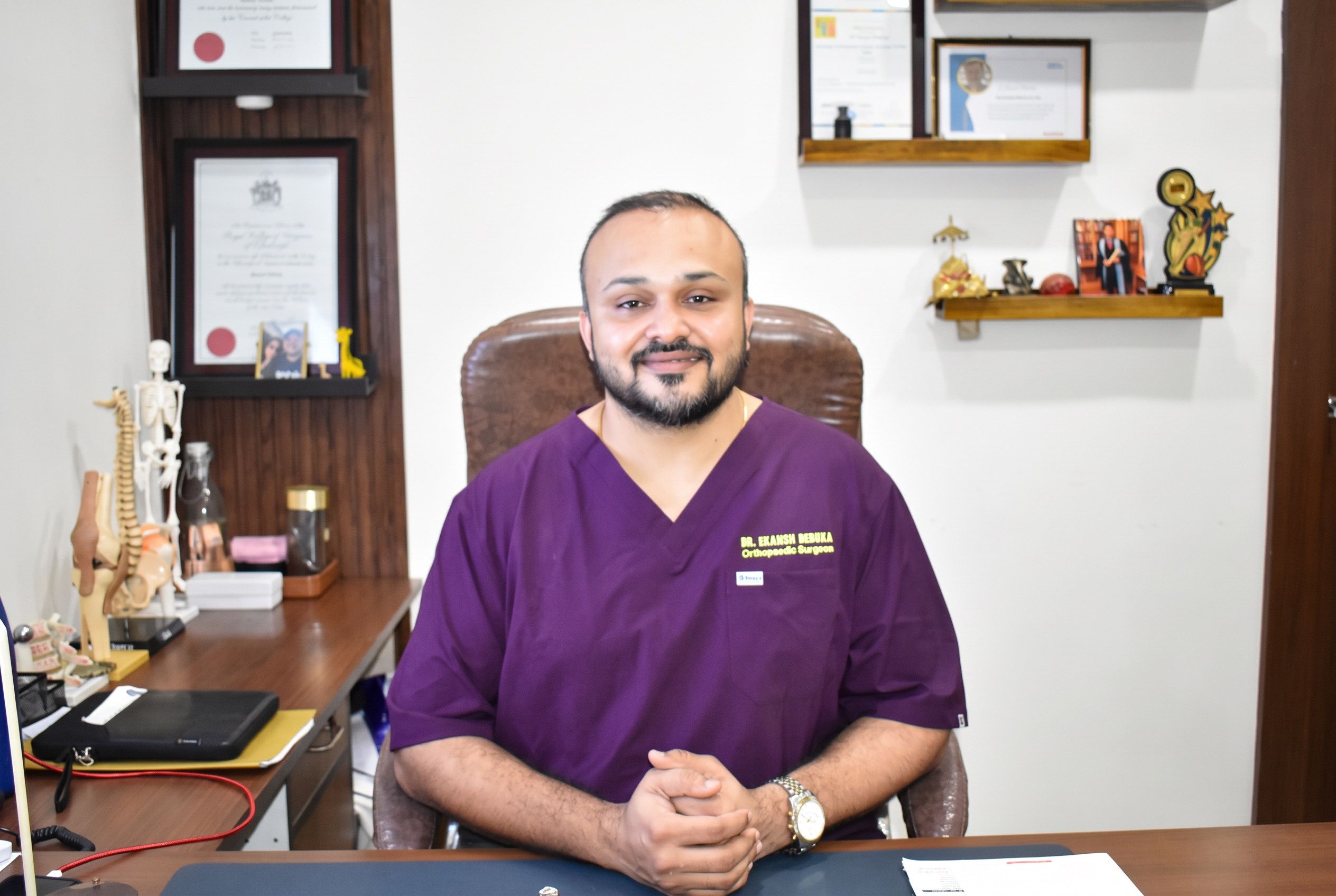 Dr. Ekansh Debuka at his clinic desk in Ranchi, with spine and joint models and framed qualifications